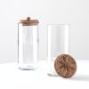 Two tall clear glass jars with carved wooden lids, one closed and one open with its lid leaning against it.